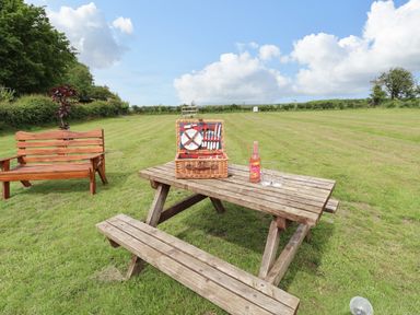 A wooden picnic table with a basket of cutlery a bottle and two glasses on a lawn near a wooden bench at Honeybee Lodge in Stixwould near Woodhall Spa