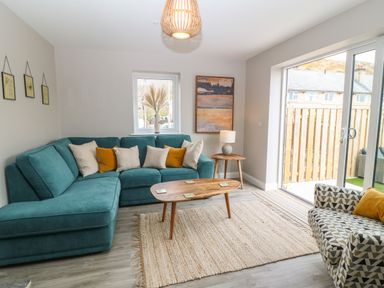 A living room with teal sectional sofa and patterned armchair near a wooden coffee table and sliding glass door at 1 Crud Y Mor in Barmouth