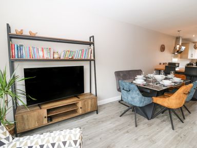 A living room with a television on a wooden stand and a dining table with chairs set with dishes at 1 Crud Y Mor in Barmouth