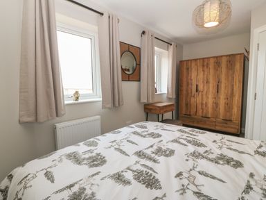 A bedroom with a bed covered in patterned bedding wooden wardrobe small side table and two windows with curtains at 1 Crud Y Mor in Barmouth