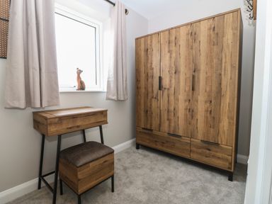 A room with a wooden wardrobe a small wooden desk with a padded stool a window with curtains and a wooden cat figurine on the windowsill at 1 Crud Y Mor in Barmouth