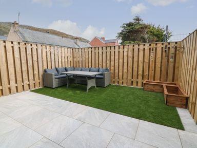 A fenced outdoor patio with artificial grass a wicker corner sofa a table and wooden planters at 1 Crud Y Mor in Barmouth