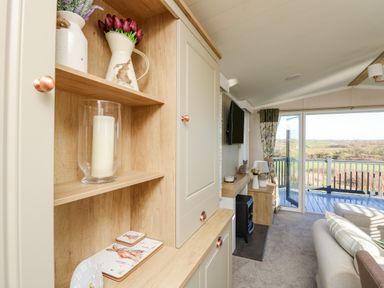 A living room with shelves holding a candle and flower vases a television on the wall a sofa and glass doors opening to a balcony overlooking fields at Vendee Lodge Thorness Bay Holiday Park near Cowes