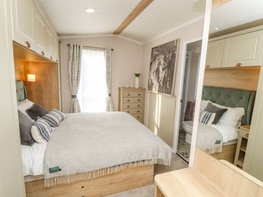 A bedroom with a double bed, overhead cabinets, a chest of drawers, window with curtains, and an open door leading to a bathroom at Vendee Lodge Thorness Bay Holiday Park near Cowes