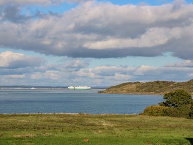 A view of a grassy field leading to a body of water with a ship and hills under a partly cloudy sky at Vendee Lodge Thorness Bay Holiday Park near Cowes