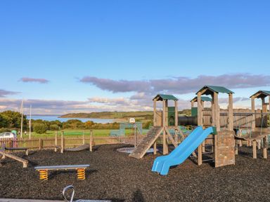 An outdoor playground with slides climbing structures and swings near a grassy field and water at Vendee Lodge Thorness Bay Holiday Park near Cowes