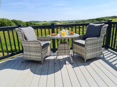 A balcony with two cushioned wicker chairs and a round table set with breakfast near a railing with countryside view at Vendee Lodge Thorness Bay Holiday Park near Cowes
