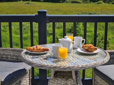 A round glass table with two croissants on plates two cups a teapot and two glasses of orange juice on a balcony with a green field view at Vendee Lodge Thorness Bay Holiday Park near Cowes