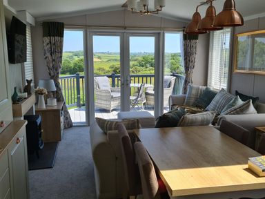 A living room with sofa and cushions a wooden dining table and chairs and glass doors leading to a balcony with outdoor seating at Vendee Lodge Thorness Bay Holiday Park near Cowes