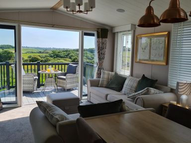 A living room with sofas and pillows adjacent to a balcony with chairs and a table overlooking green fields at Vendee Lodge Thorness Bay Holiday Park near Cowes