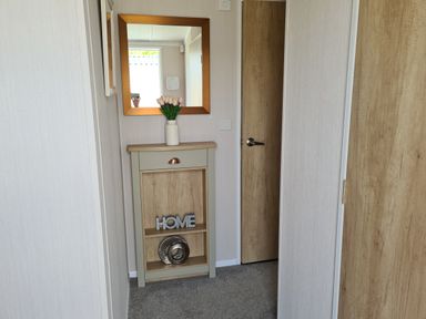 A hallway with a small cabinet holding a vase of flowers and decorative items with a square mirror hanging above at Vendee Lodge Thorness Bay Holiday Park near Cowes
