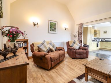 A living room with two brown armchairs and a loveseat facing a wooden coffee table with a kitchen visible in the background at Caerffynon Bach in Dyffryn Ardudwy