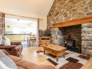 A living room with a stone fireplace a wooden coffee table a television and a brown sofa adjacent to a kitchen at Caerffynon Bach in Dyffryn Ardudwy