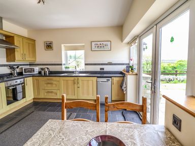 A kitchen with wooden cabinets a dining table and chairs and glass doors leading outside at Caerffynon Bach in Dyffryn Ardudwy