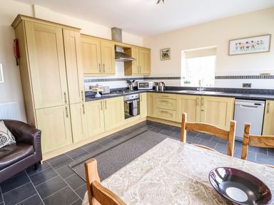 A kitchen with wooden cabinets a dining table and chairs a leather armchair and kitchen appliances at Caerffynon Bach in Dyffryn Ardudwy