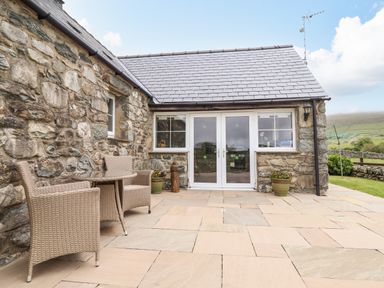 An outdoor patio with stone walls wicker chairs a small round table potted plants and glass double doors at Caerffynon Bach in Dyffryn Ardudwy