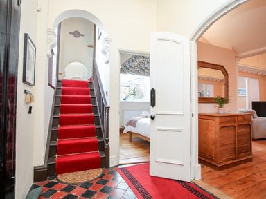 An entryway with a staircase covered by a red carpet a bedroom visible through a doorway and a wooden cabinet with a mirror in a living room at Ty Biwmares in Beaumaris
