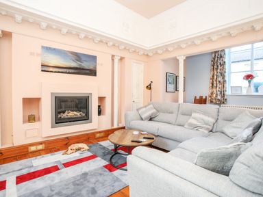 A living room with a gray sectional sofa a round wooden coffee table a fireplace and a large window with floral curtains at Ty Biwmares in Beaumaris