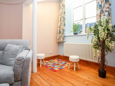 A corner of a room with a gray sofa two small stools a colorful play mat a radiator and potted plants at Ty Biwmares in Beaumaris