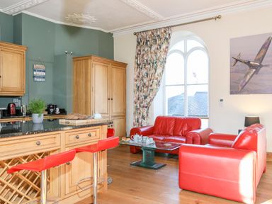 A kitchen and living area with red chairs wooden cabinets a glass coffee table and a large window at Ty Biwmares in Beaumaris