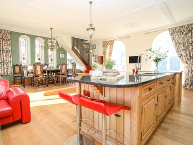 A kitchen with an island and red stools next to a dining area with wooden chairs and a chandelier at Ty Biwmares in Beaumaris