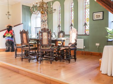 A dining area with wooden chairs and a table under a chandelier near stained glass windows and a bear statue in a colorful shirt at Ty Biwmares in Beaumaris