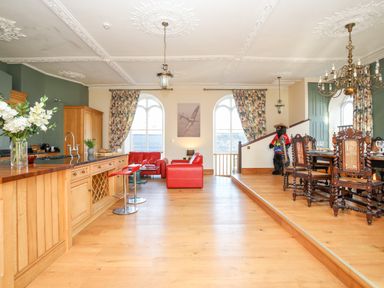 An open living and dining area with wooden floors wooden kitchen cabinets red leather sofas a dining table with chairs and floral curtains at Ty Biwmares in Beaumaris