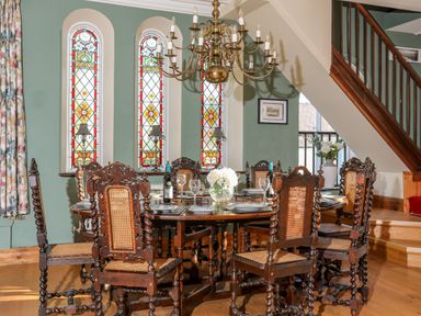 A dining room with a wooden table and chairs under a chandelier by stained glass windows at Ty Biwmares in Beaumaris
