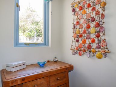 A wooden dresser under a window with a decorative box and glass items next to a wall hanging of seashells at Periwinkle Atlantic Highway near Bude