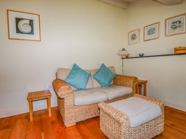 A seating area with a two-seater wicker sofa with cushions and matching footstool and wooden side tables at Periwinkle Atlantic Highway near Bude