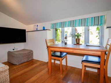 A dining area with a wooden table, two chairs, wine bottle, glasses, and a plant by a window at Periwinkle Atlantic Highway near Bude