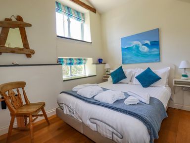 A bedroom with a double bed blue pillows waves painting wooden chair and two windows at Periwinkle Atlantic Highway near Bude