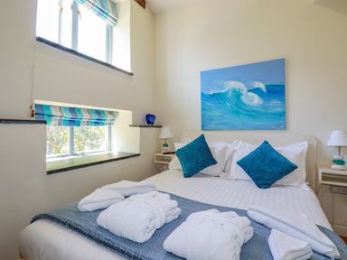 A bedroom with a bed two blue pillows white robes on a blanket two windows with striped blinds and a wave painting above the bed at Periwinkle Atlantic Highway near Bude