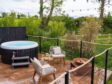 An outdoor patio with a hot tub two chairs a small table with drinks and snacks and a fire pit surrounded by greenery at Rusty Boathouse in Llanarth