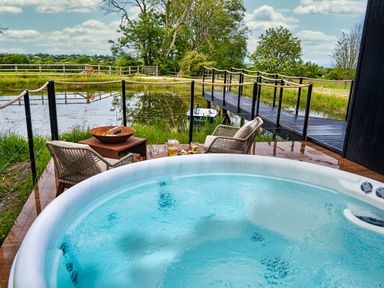 A hot tub with two chairs and a fire pit overlooking a pond with a wooden bridge at Rusty Boathouse in Llanarth