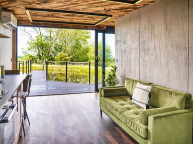 A room with a green sofa and kitchen counter with stools overlooking a deck and trees at Rusty Boathouse in Llanarth