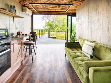 An open plan living space with a green sofa kitchen area with bar stools and a large sliding door opening to a deck overlooking water at Rusty Boathouse in Llanarth