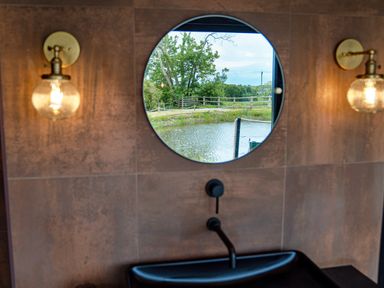 A bathroom sink with round mirror reflecting an outdoor view of a pond and trees at Rusty Boathouse in Llanarth