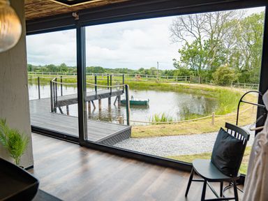 A room with a wooden floor looking out through a large window at a small pond with a dock and surrounding grass at Rusty Boathouse in Llanarth