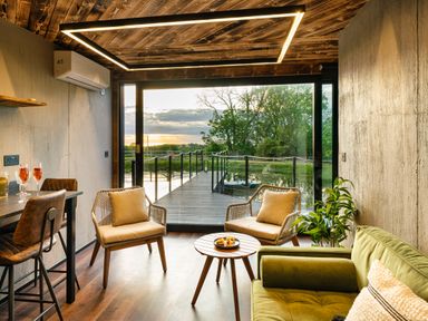 A sitting area with chairs a green sofa and a small table overlooking a wooden deck and water at Rusty Boathouse in Llanarth