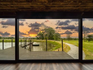 A view of a pond with a wooden deck and rope railing outside large windows with a sunset and gravel path at Rusty Boathouse in Llanarth