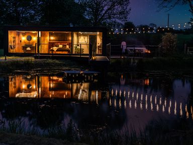 A modern cabin with a bedroom and bathroom lit up at night by a lake with string lights and outdoor seating at Rusty Boathouse in Llanarth