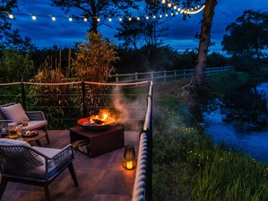 An outdoor patio with chairs a fire pit string lights and a lantern by a river at Rusty Boathouse in Llanarth