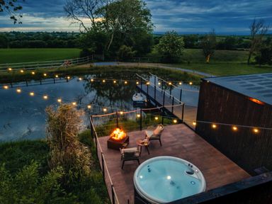 A patio with a hot tub fire pit and chairs next to a pond with string lights at Rusty Boathouse in Llanarth