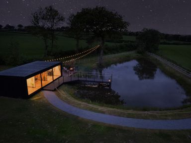 A modern cabin with lights on near a pond with a wooden deck and string lights at Rusty Boathouse in Llanarth