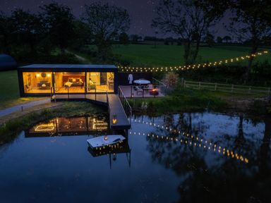 A modern cabin with large windows overlooking a pond with a dock and boat at Rusty Boathouse in Llanarth