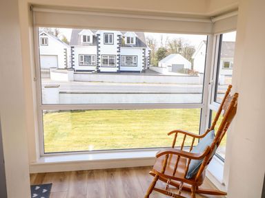 A wooden rocking chair with a cushion near a large window showing a grassy yard and white houses across the street at Bridgetown Cottage in Kerrykeel County Donegal