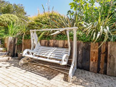A white metal garden swing on a paved patio with wooden planter and leafy plants at Cameo Atlantic Highway near Bude