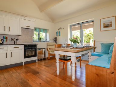 A kitchen and dining area with a wooden floor a white table with chairs a bench with blue cushions and kitchen cabinets at Cameo Atlantic Highway near Bude
