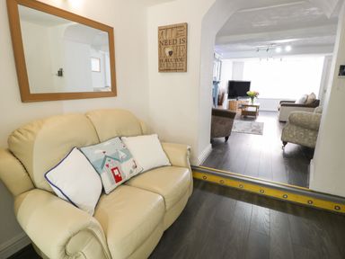 A beige leather loveseat with three pillows and a mirror on the wall next to an open archway leading to a living room with a television a coffee table and armchairs at Penrallt in Cemaes Bay